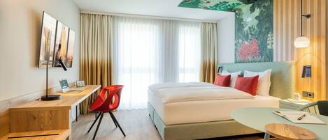 A hotel room with a bed, a desk with a computer, a chair, a small table, and a painting on the wall.