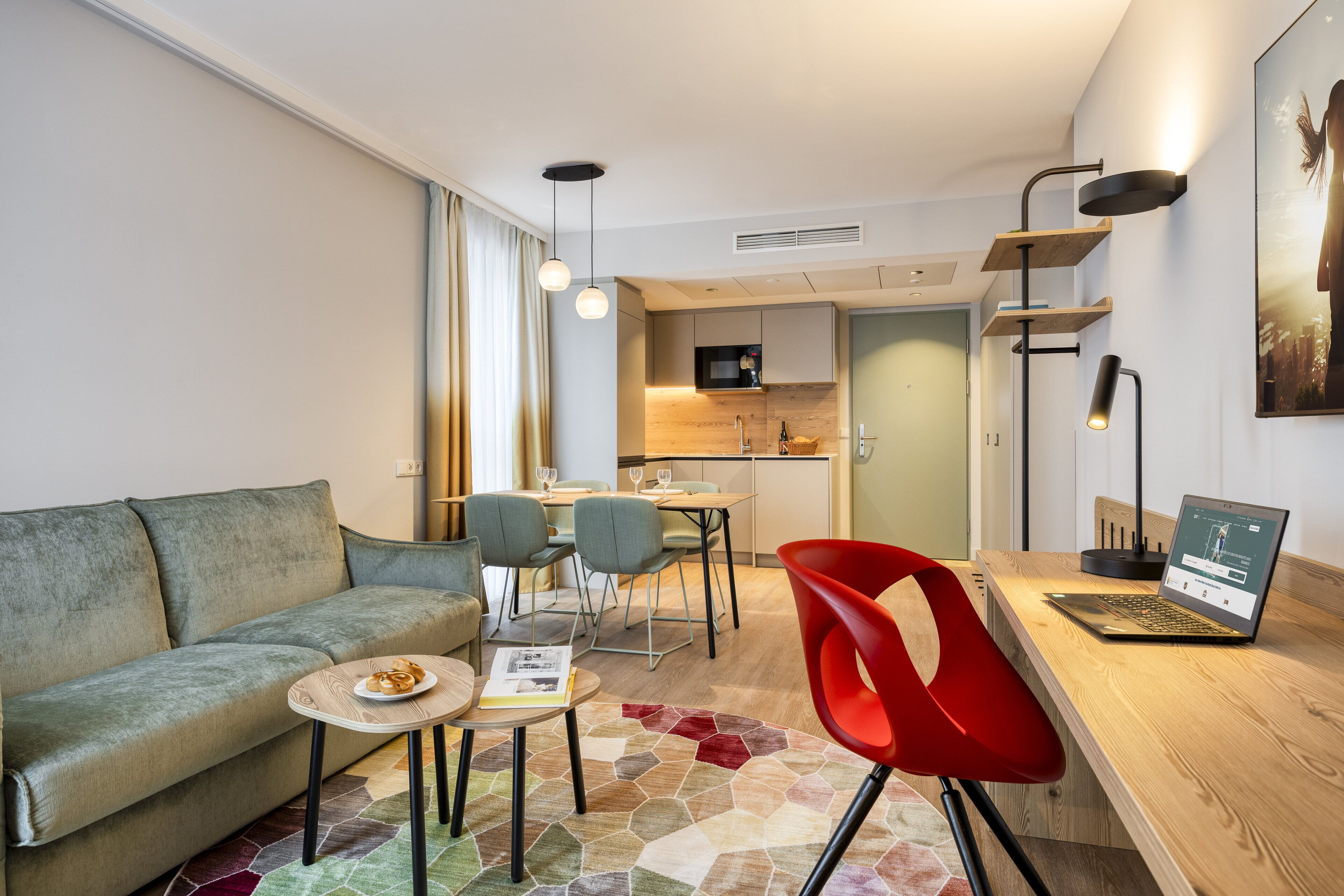 A modern living room with a sofa, dining table, and chairs, a kitchen area with a microwave, and a laptop on a wooden desk.