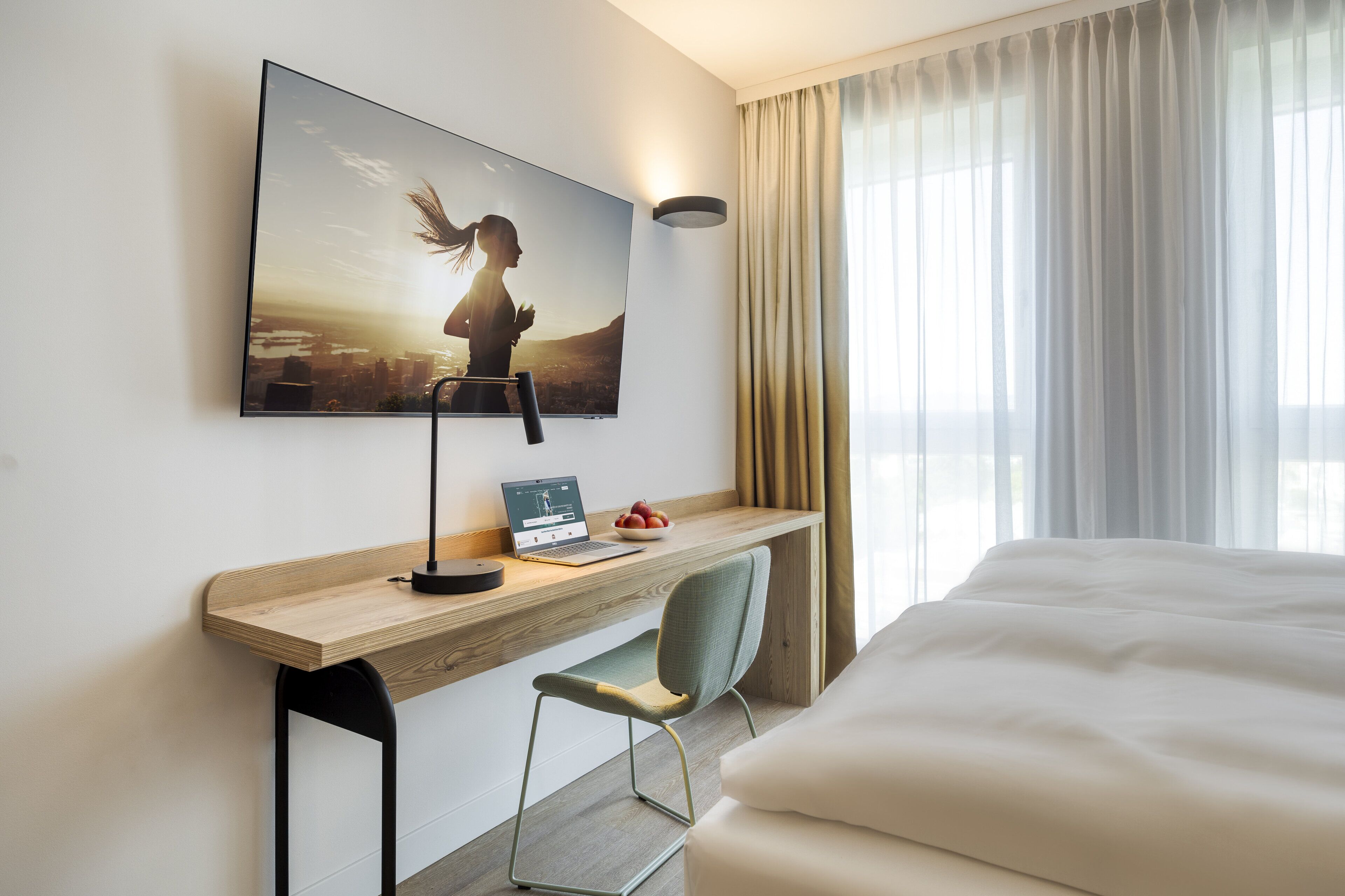 Een hotelkamer met een bed, een bureau met een laptop, een stoel, een aan de muur bevestigde televisie en een raam met gordijnen.