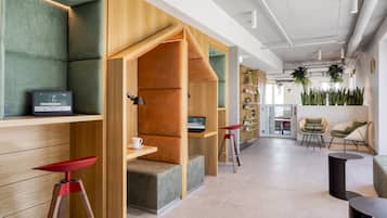 Un espacio de oficina moderno con zonas de estar con asientos de madera y acolchados en tonos verdes, un ordenador portátil sobre una estantería y vistas al área de comedor.
