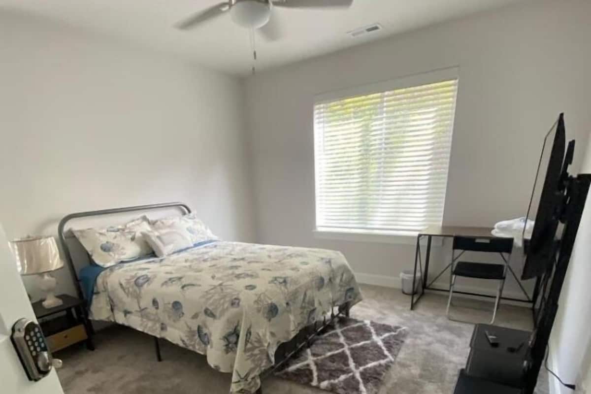 A bedroom with a bed, a desk, a chair, a ceiling fan, and a window with blinds.
