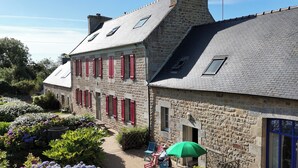 A stone building with red shutters, a garden with hydrangeas, and a patio with outdoor seating. - NEW: La Grange (Ploubezre)