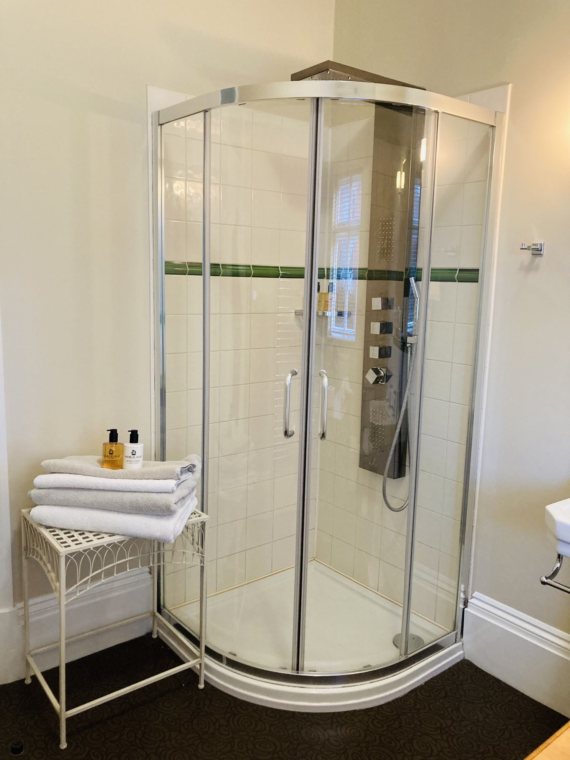 A glass-enclosed shower stall with a white tiled wall, a towel rack with folded towels, and a small shelf with toiletries.