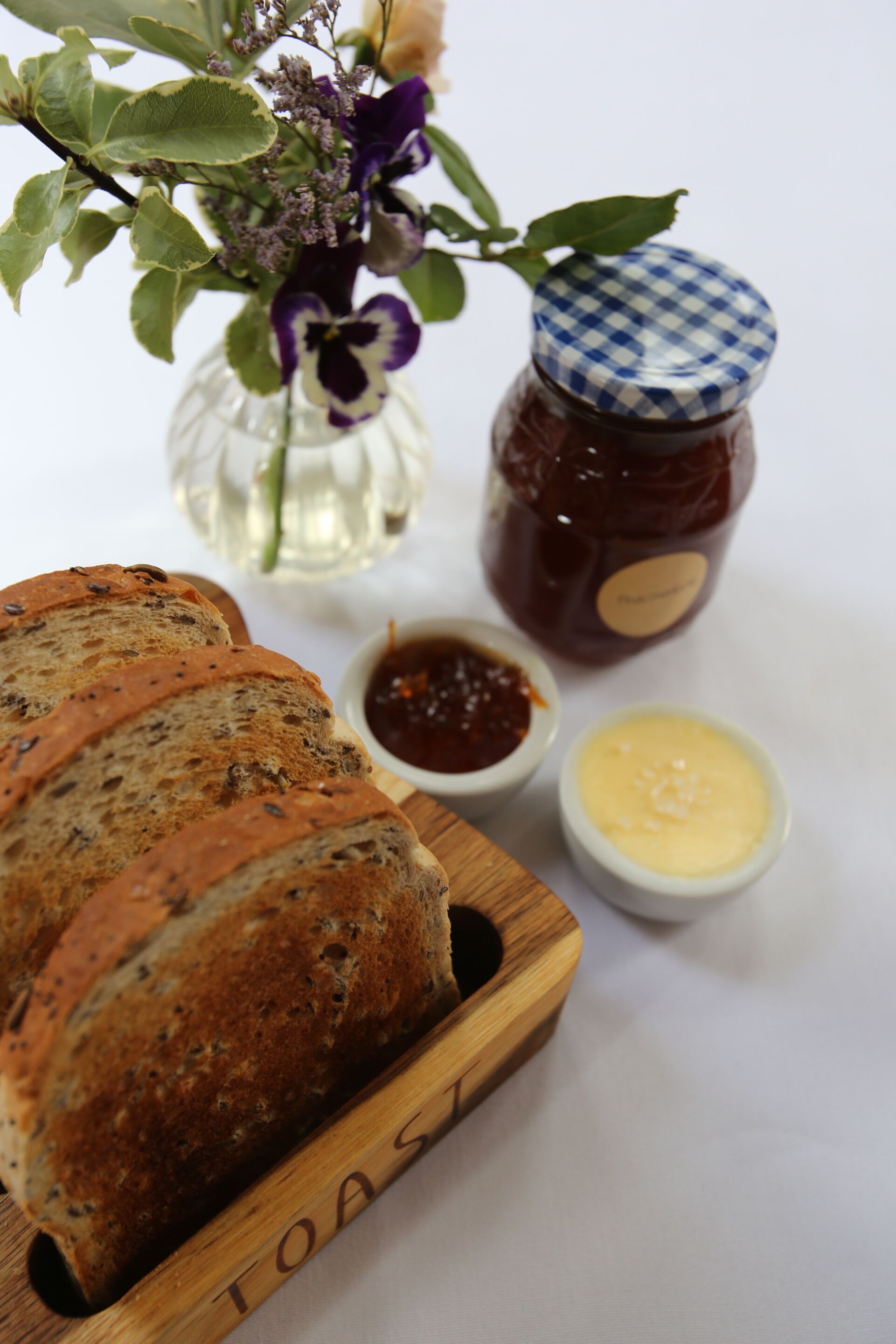 A wooden board with sliced bread, a jar of jam, and small bowls of spreads.
