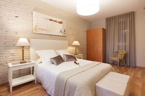 Um quarto com parede de tijolos brancos, guarda-roupa de madeira, cama branca com manta texturizada, duas mesas de cabeceira com luminárias e uma cadeira de vime.