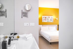 Habitación de hotel con una cama blanca, una mesita de noche con artículos de aseo, un espejo y una lámpara fijada a la pared.