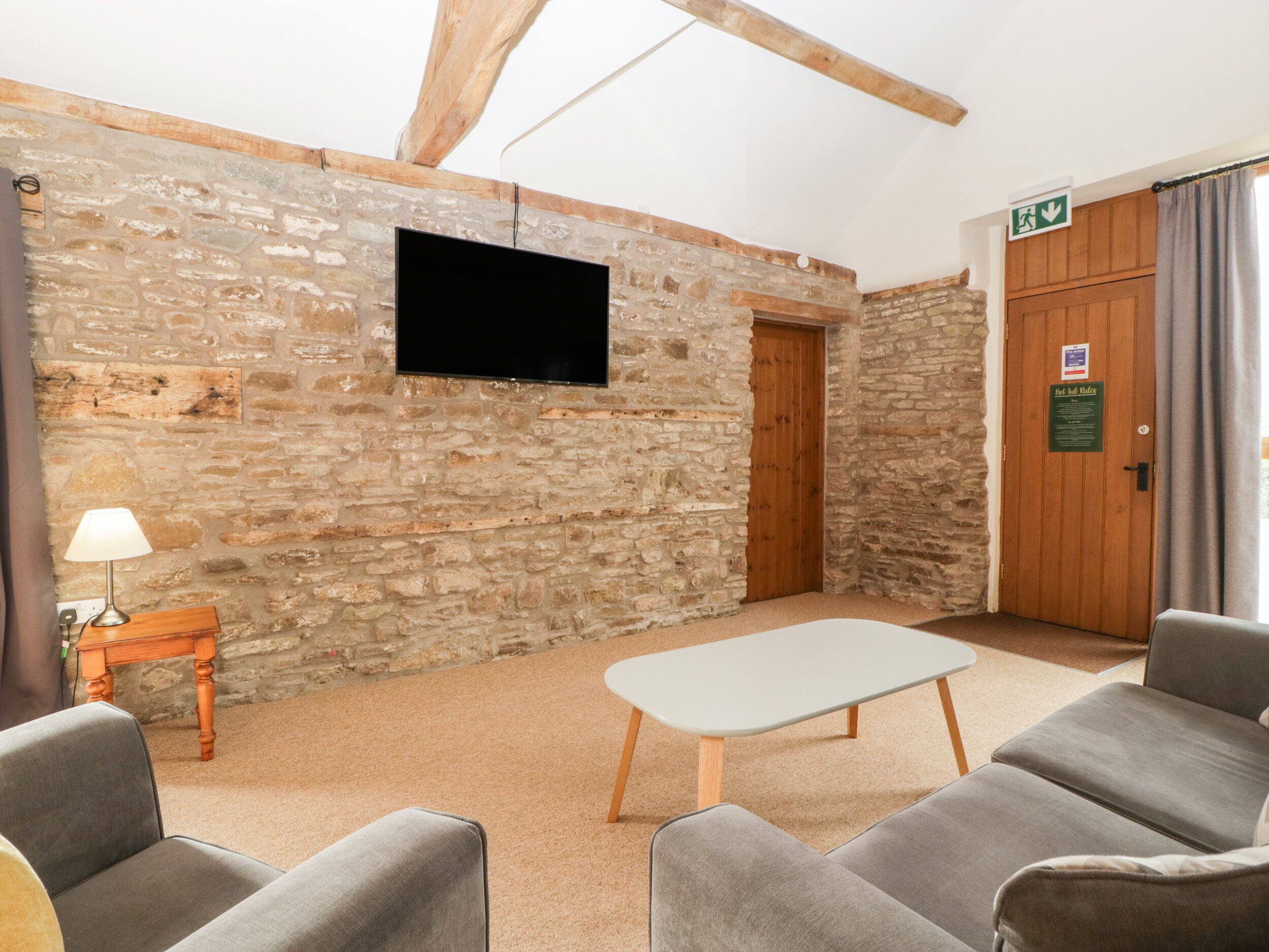 Una habitación con paredes de piedra, un televisor de pantalla plana montado en la pared, una puerta de madera y una mesa con dos sofás grises.