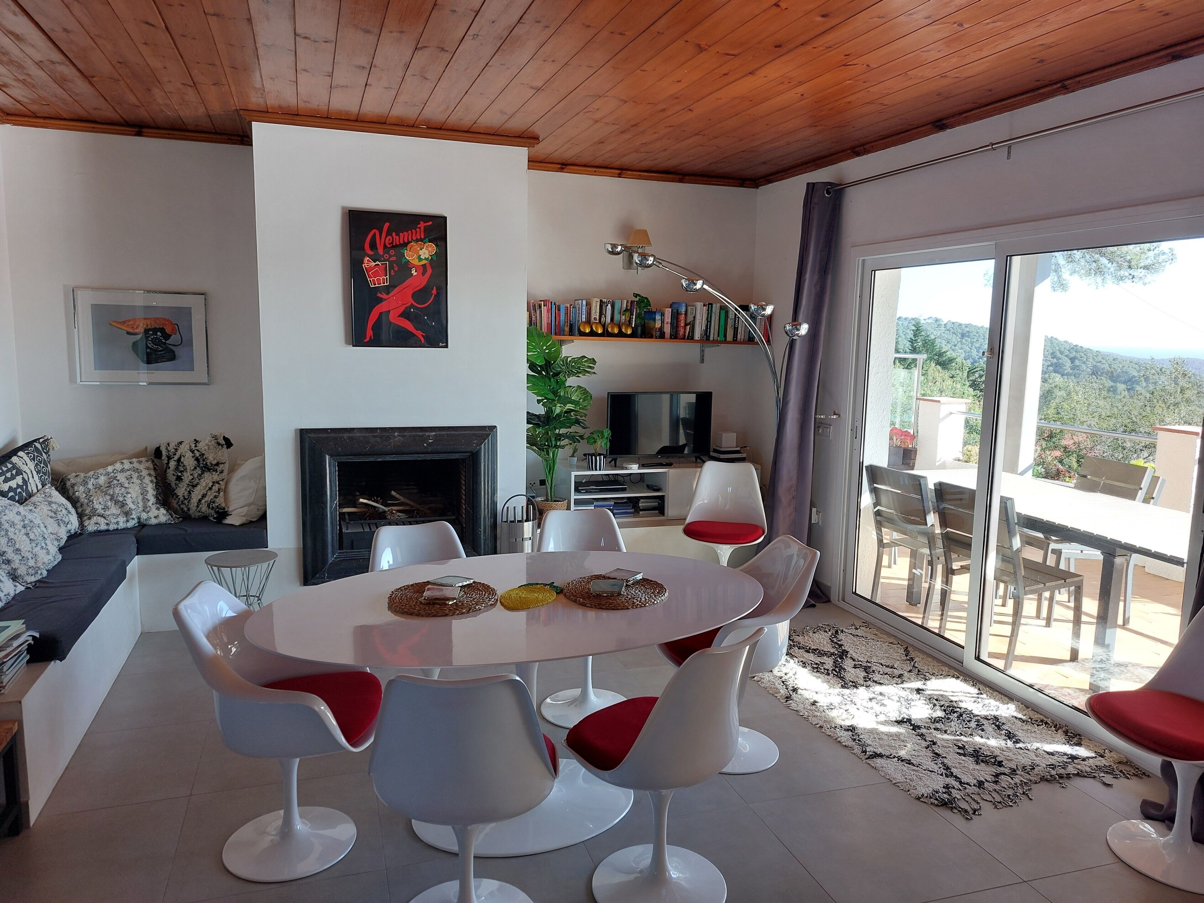A modern living room with a fireplace, a dining area with white chairs, a sofa with patterned cushions, and a bookshelf.