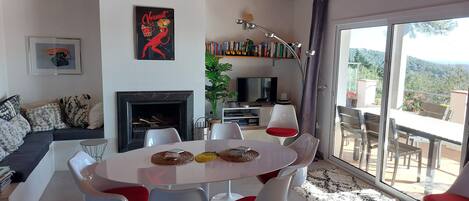 A modern living room with a fireplace, a dining area with white chairs, a sofa with patterned cushions, and a bookshelf.