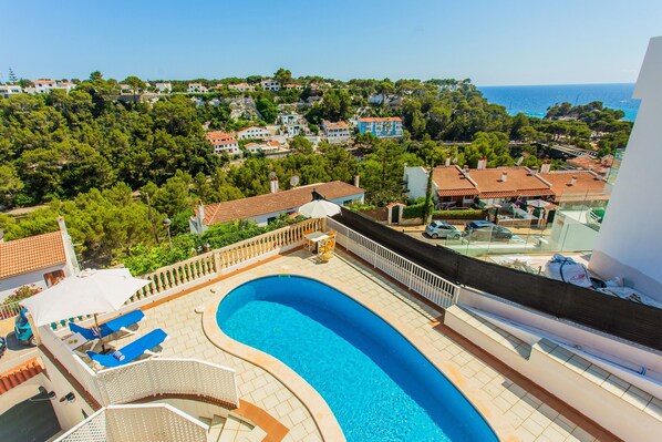 A rooftop pool with lounge chairs, a view of the sea, and a residential area in the background.