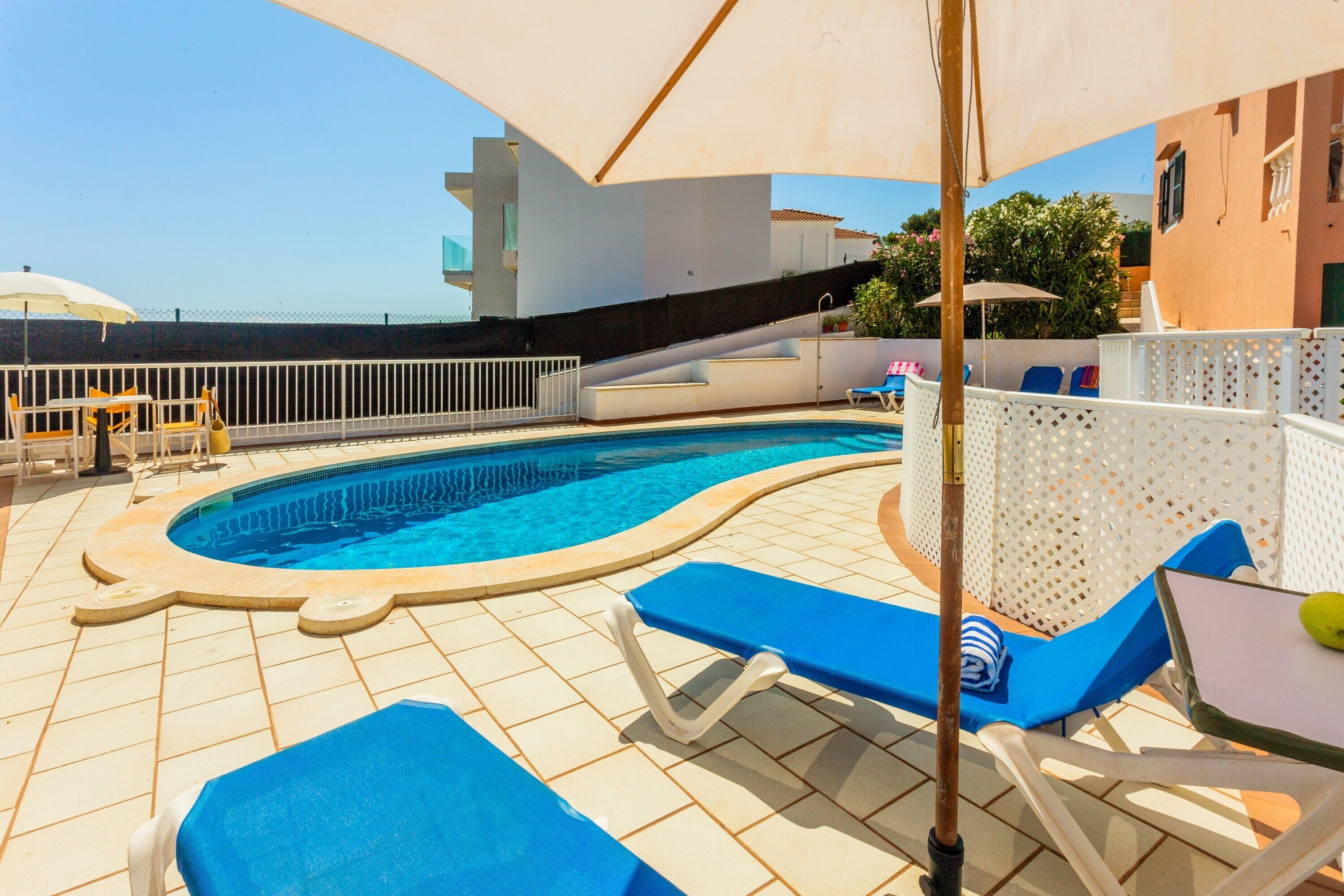 Una piscina con sillas reclinables azules, una sombrilla blanca y una valla blanca.