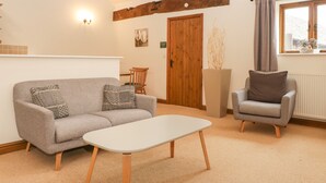 Una sala de estar con sofá, sillón y mesa de centro, vigas de madera en el techo, una puerta y una ventana con cortinas.