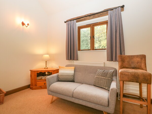 A cozy room with a grey sofa, a wooden nightstand with a lamp, a window with curtains, and a wooden chair.