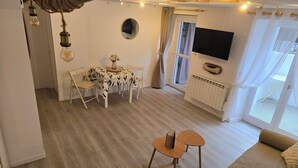 Ett mysigt vardagsrum med en soffa, ett soffbord och en matplats med ett bord och stolar. En tv är monterad på väggen, och det finns ett fönster med gardiner.