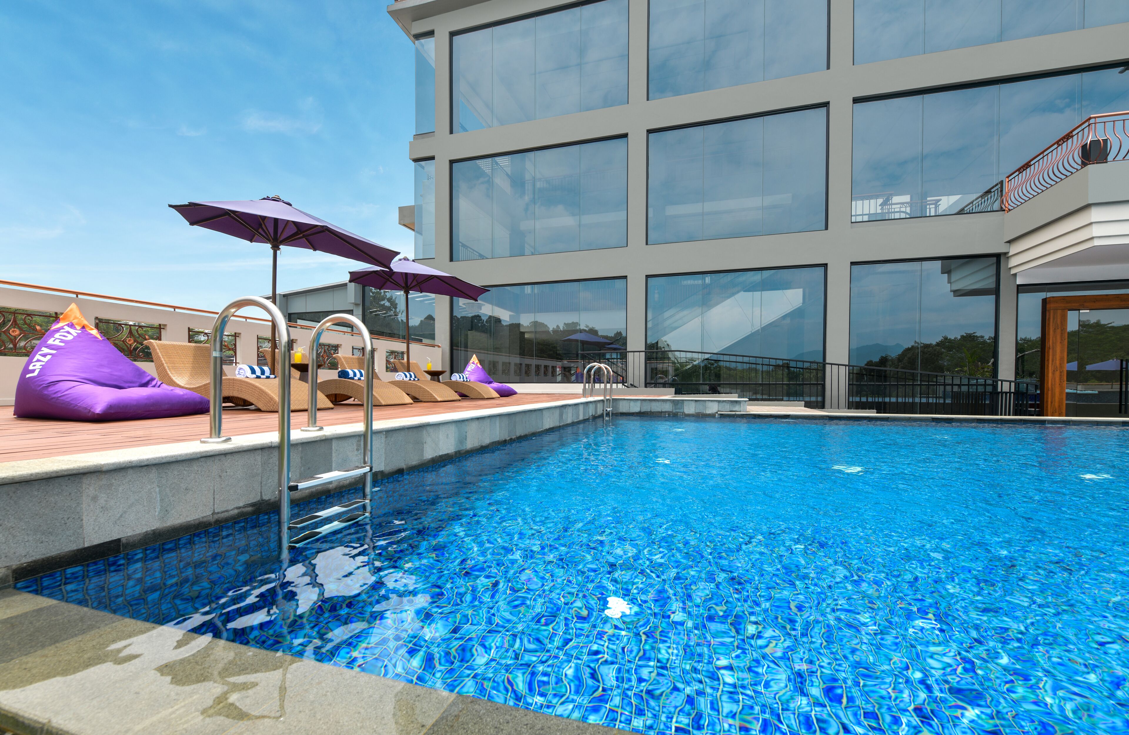 Piscina interior moderna con agua azul cristalina, rodeada de sillones de descanso y sombrillas, con un gran edificio de cristal al fondo.