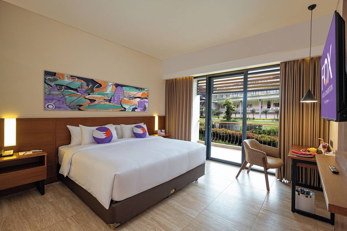 Ein Hotelzimmer mit einem großen Bett, einem Schreibtisch mit Stuhl, einem Fernseher und einem Balkon mit Vorhängen.