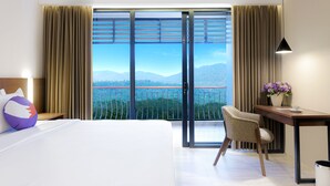 A hotel room with a bed, a desk, a chair, a nightstand, a lamp, a window with curtains, and a view of a bridge and mountains. - FOX Harris Hotel & Conventions Banjarnegara (Banjarnegara)