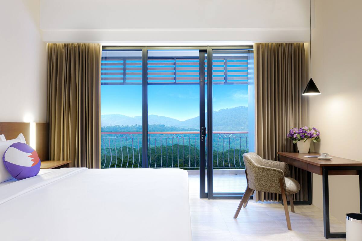 Ein Hotelzimmer mit Bett, Schreibtisch, Stuhl, Nachttisch, Lampe, einem Fenster mit Vorhängen und Blick auf eine Brücke und Berge.