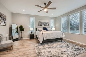 A bedroom with a bed, a ceiling fan, a large window with blinds, a mirror, and a plant.