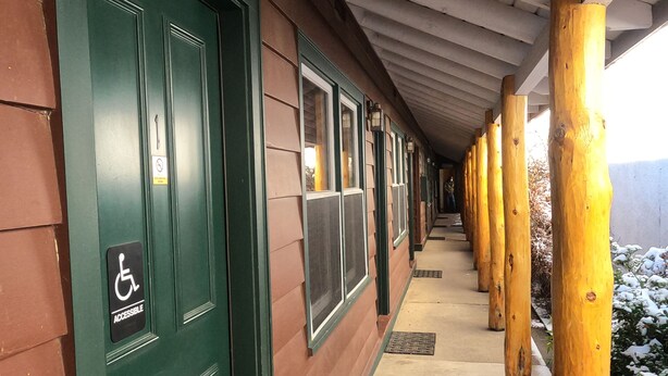 Un corredor cubierto con pilares de madera, una puerta verde con el símbolo de accesibilidad y ventanas a lo largo del lateral.