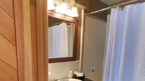 Un baño con tocador de madera, un lavamanos blanco, un espejo con iluminación, cortina de ducha y un estante pequeño con artículos de aseo.
