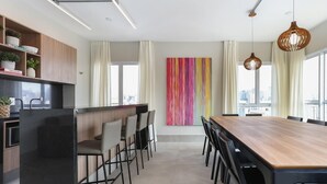 A modern kitchen with a black refrigerator, wooden cabinets, and a black countertop. There is a dining area with a wooden table and black chairs. A colorful abstract painting is on the wall, and there are large windows with curtains. - Studio Moderno Bela Vista | Wifi 250 mb (São Paulo)