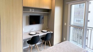 A modern bedroom with a large bed, a desk with two chairs, a TV, and a balcony with a view. - Studio Moderno Bela Vista | Wifi 250 mb (São Paulo)