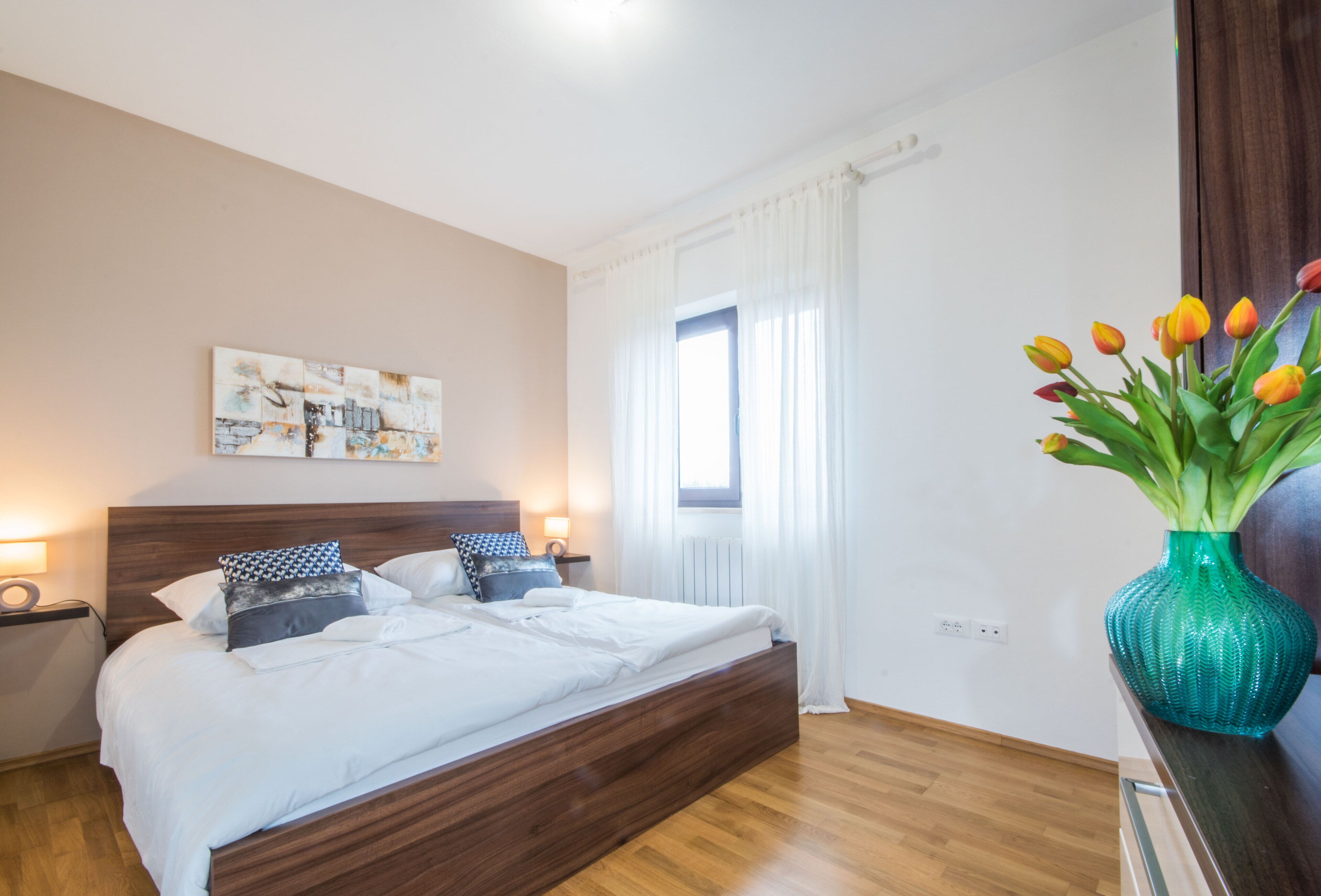 A bedroom with a wooden bed, white bedding, a wall-mounted artwork, a window with curtains, and a vase with tulips on a nightstand.