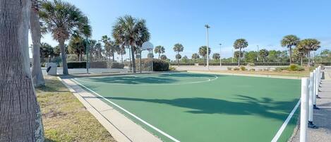 Una cancha de baloncesto al aire libre con superficie verde, rodeada de árboles y una valla.