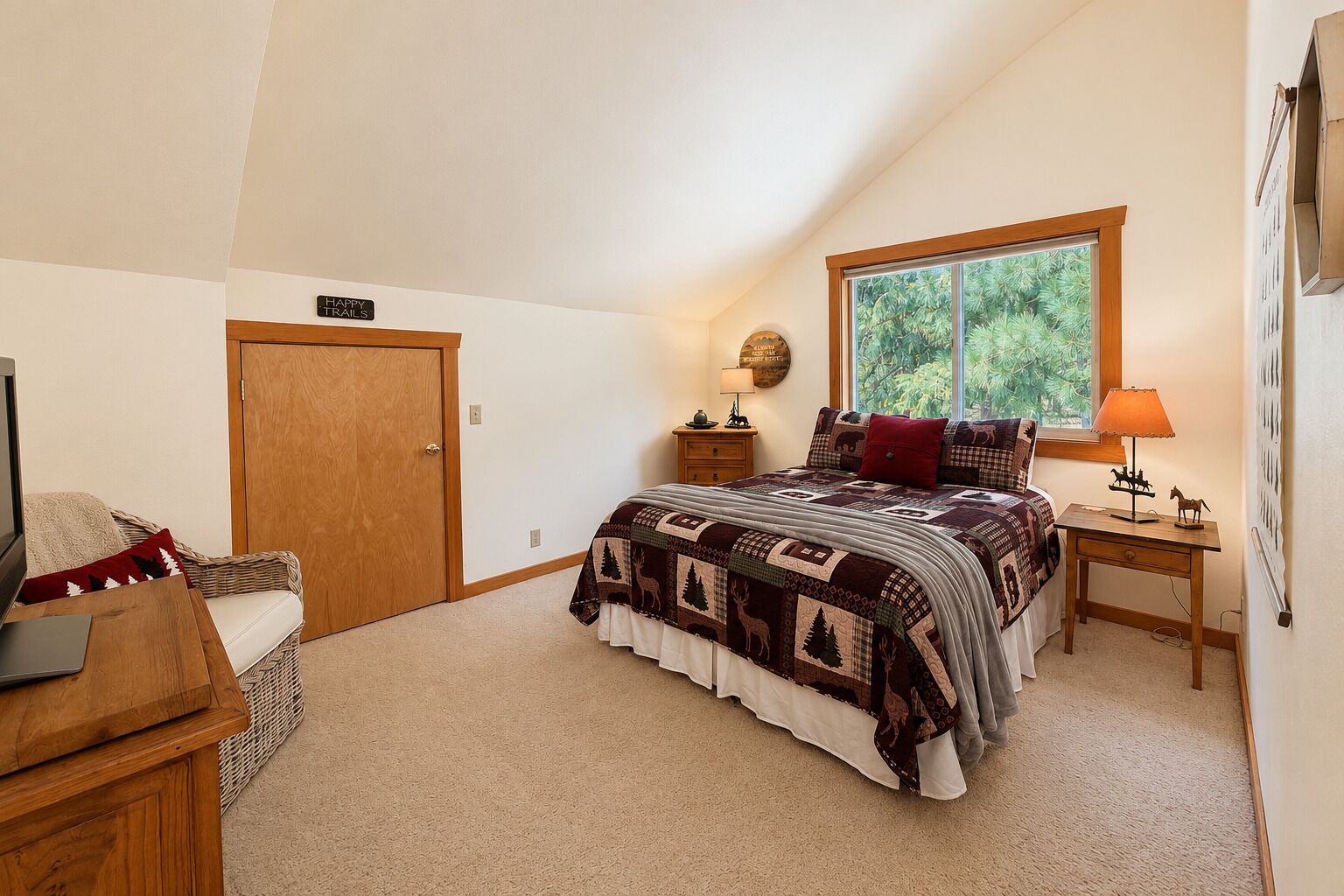 Een slaapkamer met een schuine plafond, een bed met een bedsprei met patroon, een nachtkastje met een lamp, een houten dressoir en een raam met uitzicht op groen.