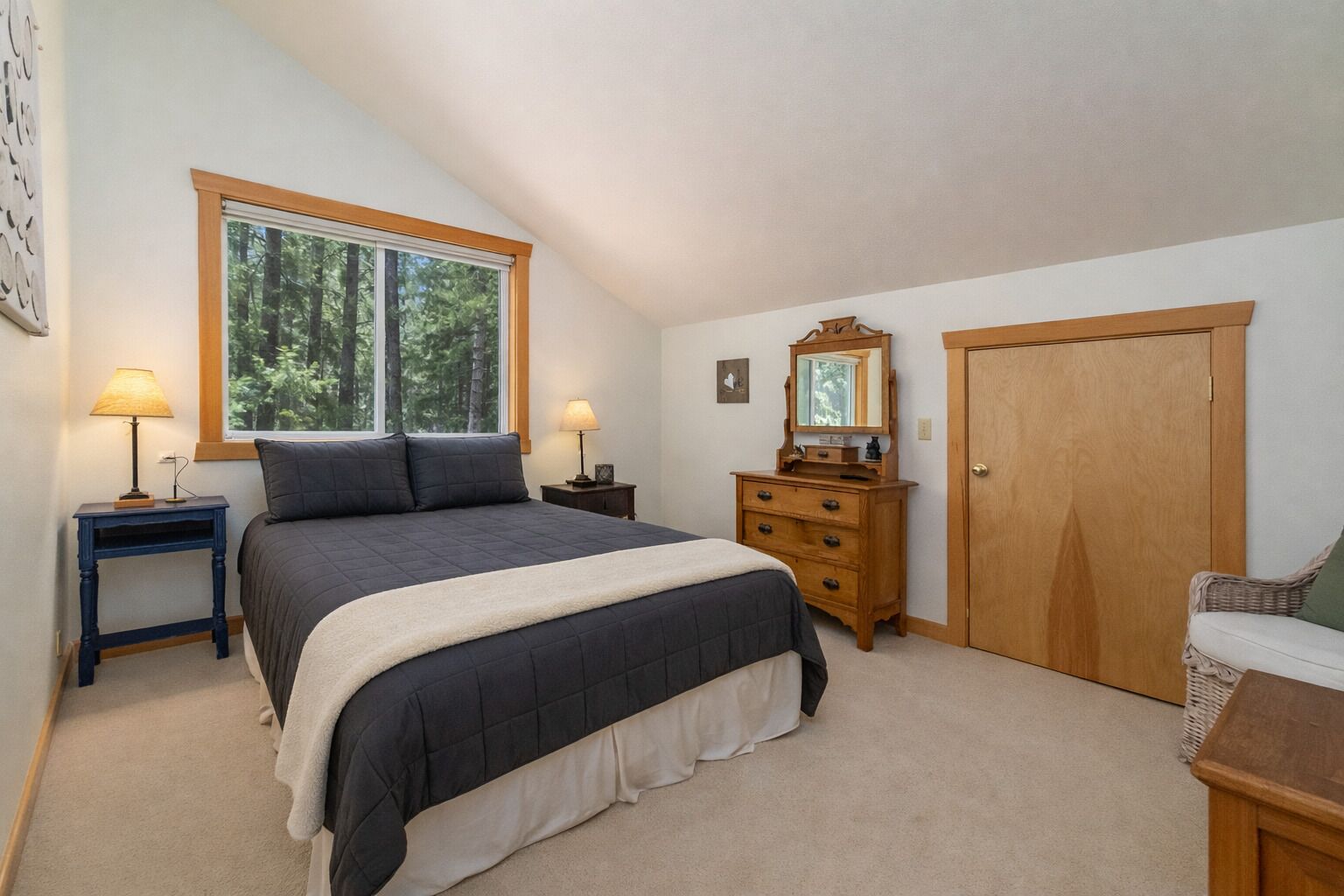 Une chambre à coucher avec un grand lit, deux tables de chevet avec des lampes, une commode avec un miroir, une porte et une fenêtre donnant sur des arbres.