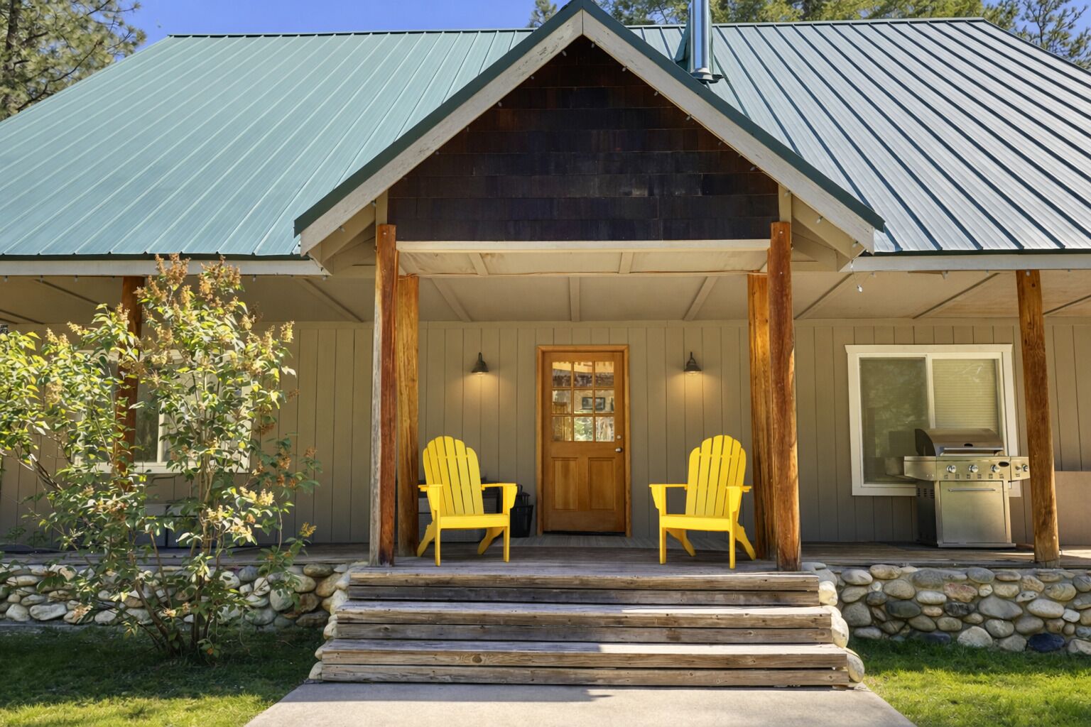 A cabin with a metal roof, wooden porch, two Adirondack chairs, a stone wall, and a grill.