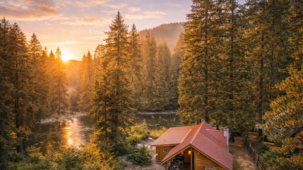A cabin with a red roof surrounded by dense forest and a river.