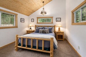 A bedroom with a wooden bed, two bedside tables with lamps, a wooden ceiling, and large windows with views of greenery.