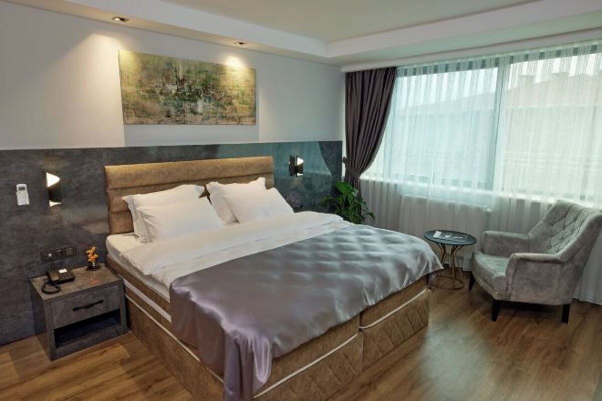 Une chambre d’hôtel moderne avec un grand lit, une table de chevet avec un téléphone, une petite table d’appoint, une chaise et une grande fenêtre avec des rideaux.