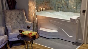 Um banheiro luxuoso com parede de mármore, uma banheira, uma poltrona estofada e uma mesinha com uma bandeja de frutas e vinho.