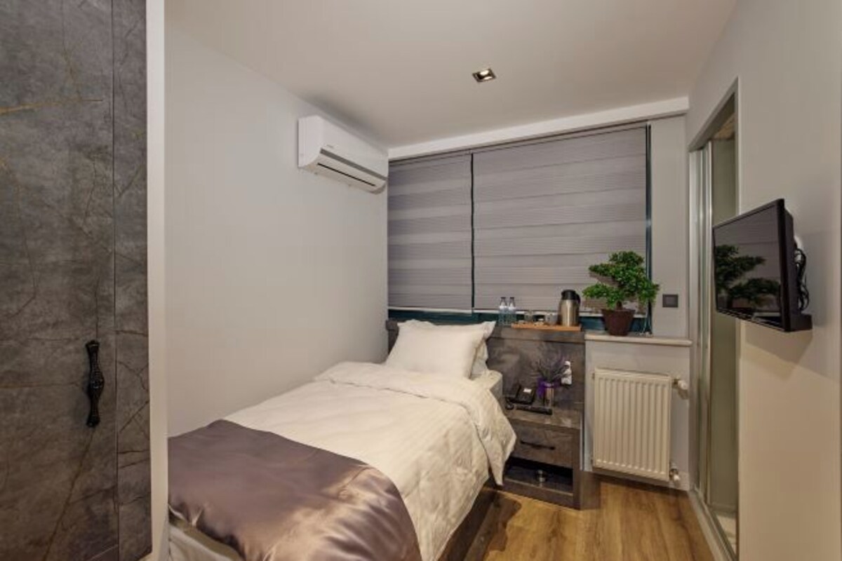 Une petite chambre à coucher moderne, équipée d’un lit, d’une table de chevet, d’un climatiseur, d’un radiateur et d’un téléviseur fixé au mur.