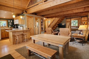 A spacious cabin interior with a wooden dining table, bar stools, a sofa, and a staircase leading to an upper level.