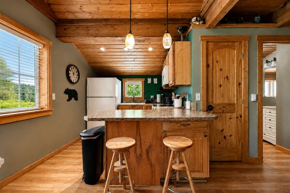 A kitchen with wooden cabinets, a central island with stools, a refrigerator, and a clock on the wall. - Lil Bigfoot Chalet 2 Bedroom Home by NW Comfy Cabins by RedAwning (Leavenworth)