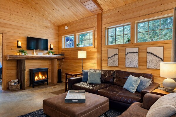 A cozy cabin interior with a fireplace, leather sofa, and large windows offering a view of the forest. - Painted Pony Cabin 2 Bedroom Home by NW Comfy Cabins by RedAwning (Leavenworth)