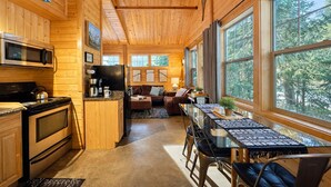 Ein gemütliches Blockhaus-Interieur mit Holzwänden, einer Küche mit modernen Geräten, einem Essbereich mit Glastisch und großen Fenstern mit Blick auf den Wald.