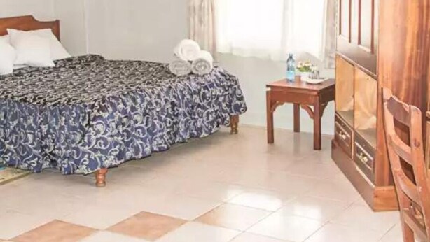 Una habitación de hotel con una cama, un armario de madera, una mesita con una lámpara y una ventana con cortinas.