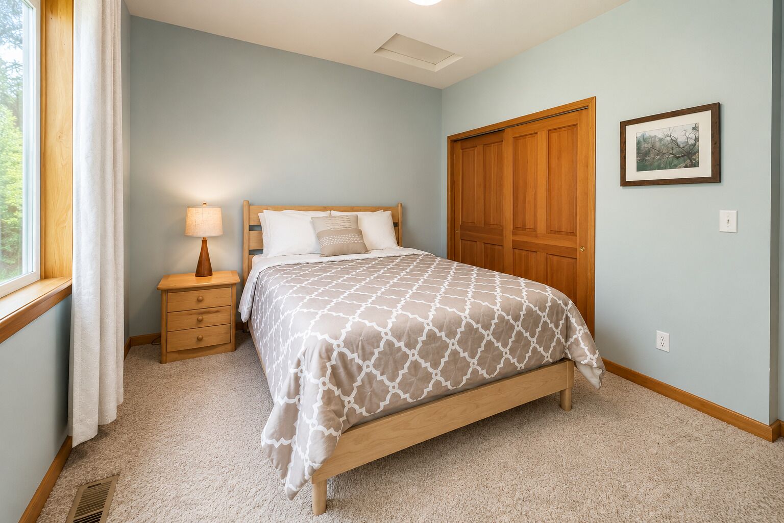 A bedroom with a bed, nightstand, lamp, wooden wardrobe, and a framed picture on the wall.