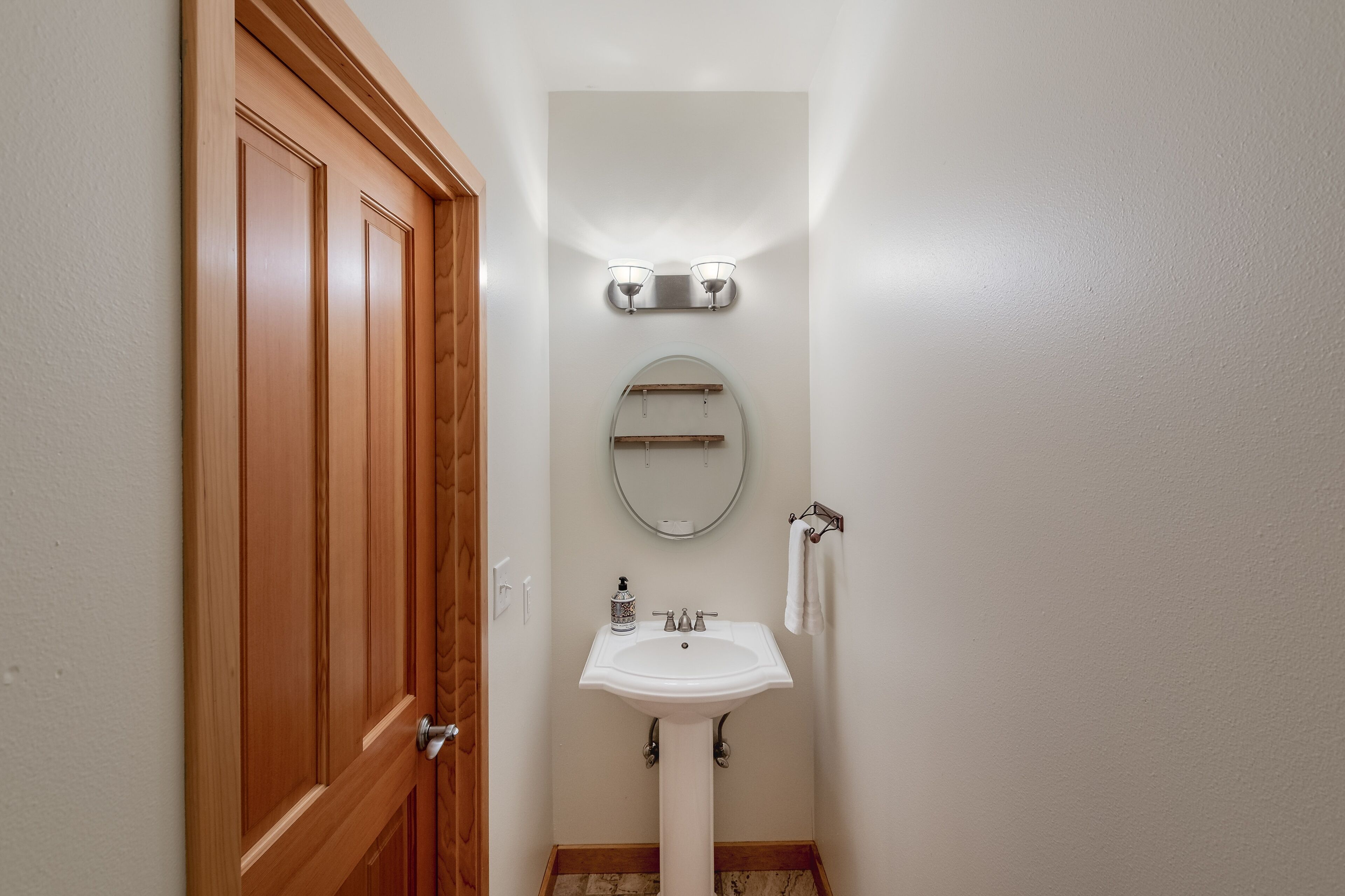 A bathroom with a wooden door, a white sink, a round mirror, and two wall-mounted lights.