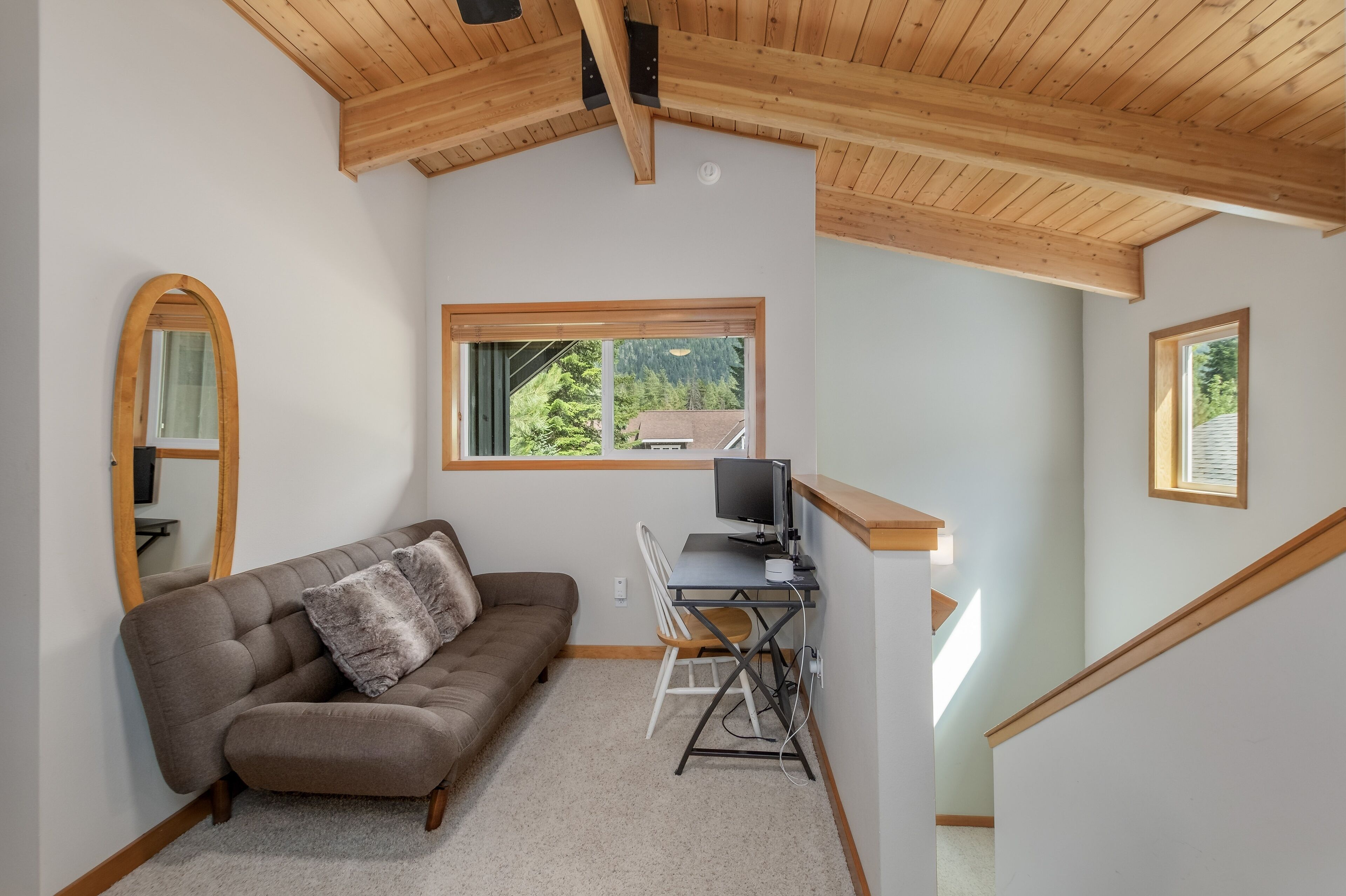 Un salon confortable avec un canapé marron, un téléviseur et un bureau avec une chaise. Il y a des fenêtres donnant sur des arbres et une maison.