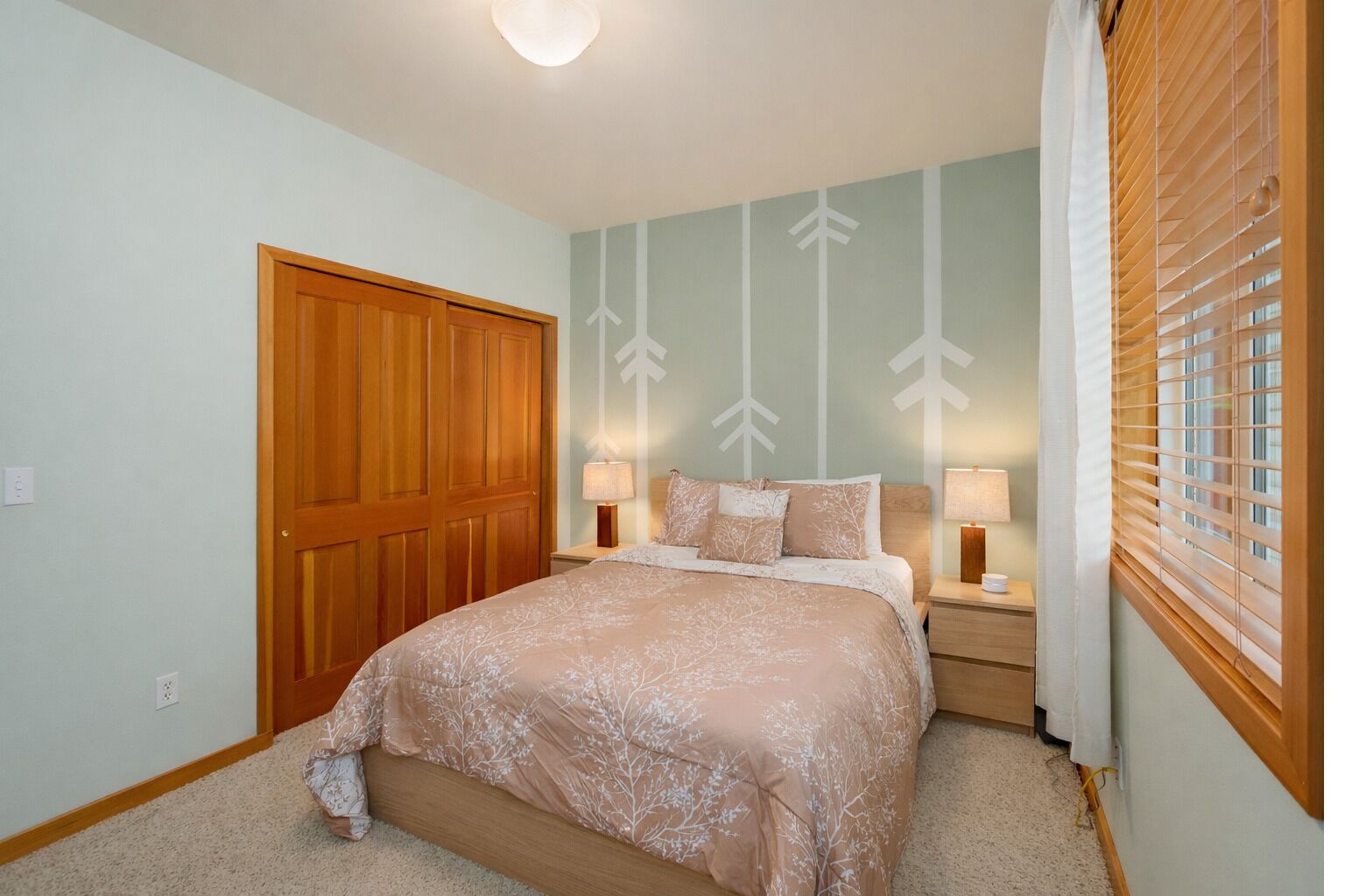 A bedroom with a large bed, two bedside tables, a wooden wardrobe, and a window with blinds.