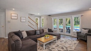 A spacious living room with a large gray sectional sofa, a wooden coffee table, and a gray armchair. There is a staircase leading to a balcony with a view of a pool and greenery. - Family Cabin 5 Bedroom Home by NW Comfy Cabins (Leavenworth)