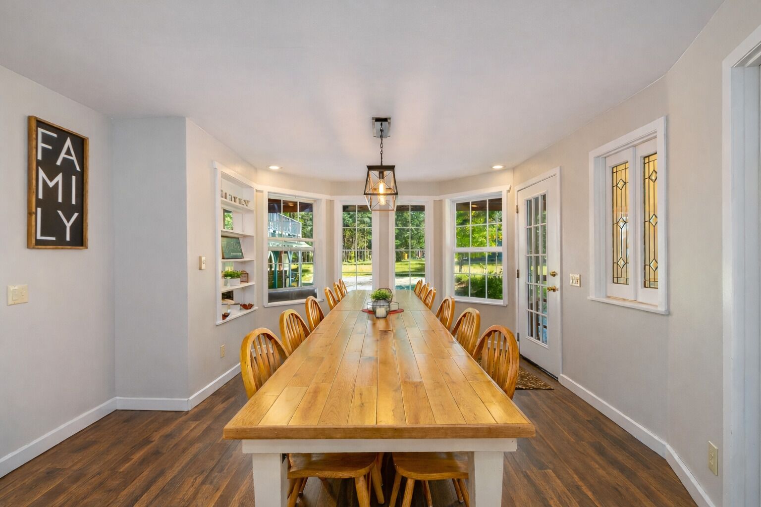 A spacious dining room with a long wooden table, surrounded by chairs, and large windows offering a view of the outdoors.