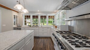 A modern kitchen with white cabinetry, a large island, a gas stove, and a view of the outdoors through large windows. - Family Cabin 5 Bedroom Home by NW Comfy Cabins (Leavenworth)