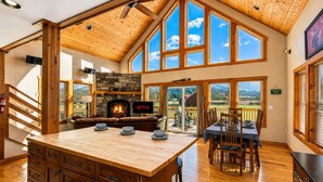 A spacious room with a fireplace, wooden ceiling, large windows, and a dining area.